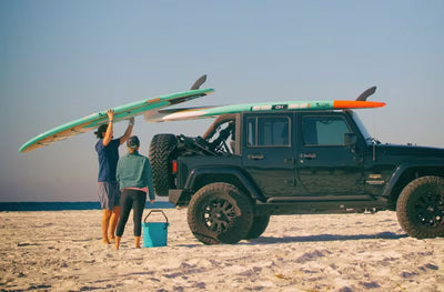 Two people loading teal Breeze Gatorshell paddle boards onto a black Jeep at the beach.