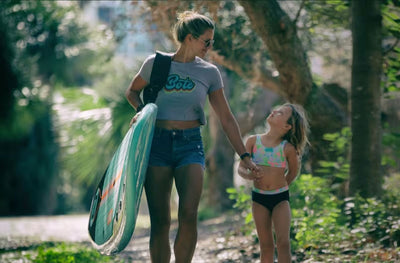 Woman carrying a Breeze Gatorshell paddle board while walking hand in hand with a child.