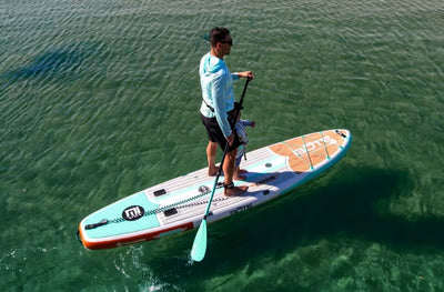 Man paddleboarding on clear water with a child seated at the front of the board.