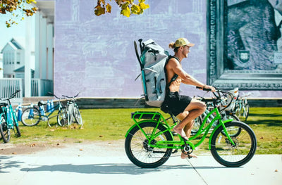 Person riding a green bike with a BOTE inflatable paddle board in a travel bag with straps on a sidewalk.