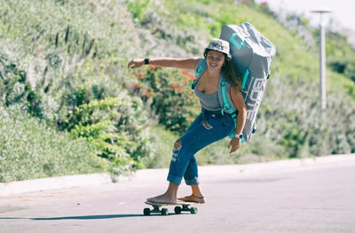Smiling woman skateboarding with a large paddle board bag strapped to her back.