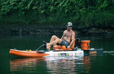 Man seated on a Rackham Aero with pedal drive system and fishing gear on calm water.