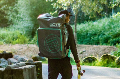 Person carrying large BOTE kayak travel bag while holding a skateboard on a wooded path.