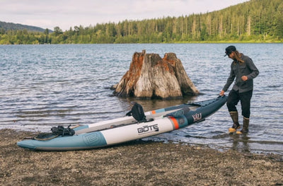 Person standing by shore pulling Vali Aero kayak onto rocky beach near a tree stump.