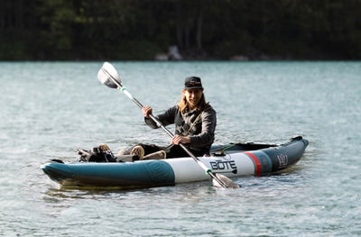 Smiling person paddling Vali Aero kayak on calm lake water.