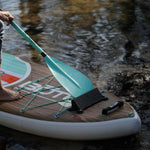 Close-up of paddle board with paddle resting on bungees near water.