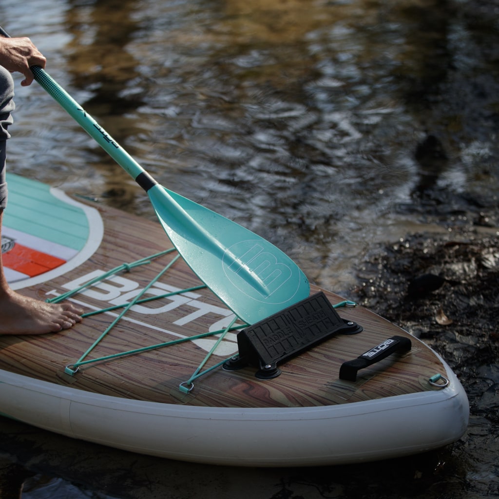 Close-up of paddle board with paddle resting on bungees near water.