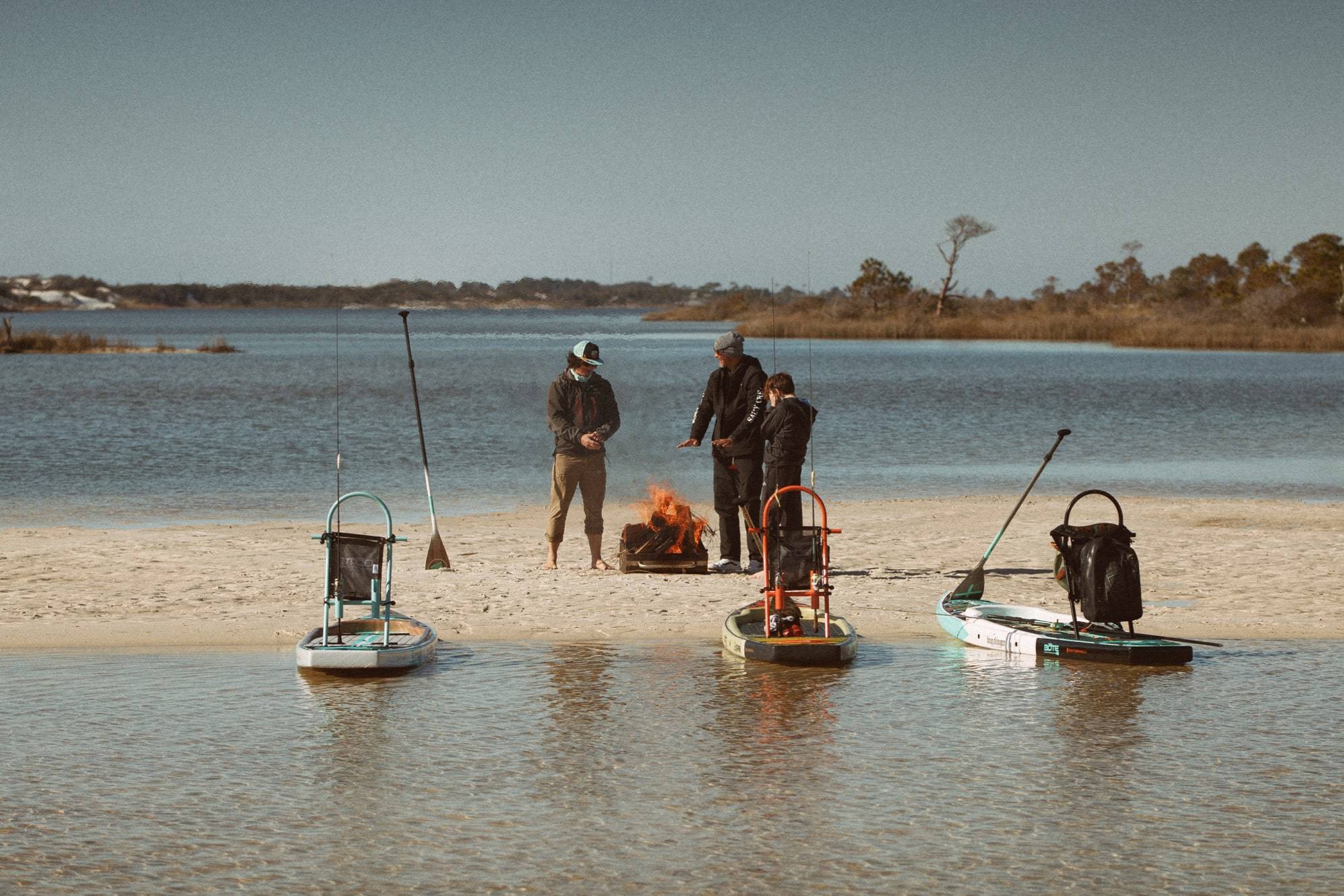 3 people standing on a sandbar around a fire warming up with their fishing paddleboards sitting in the water ready for adventure.