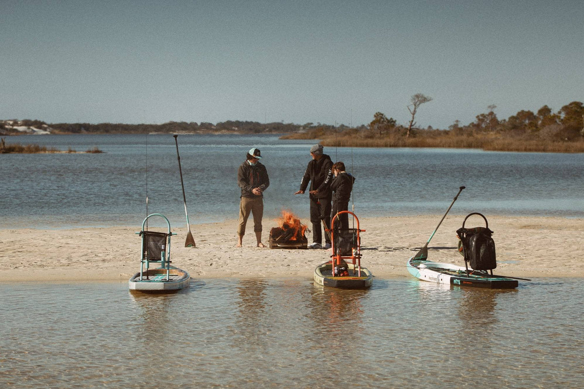 3 people standing on a sandbar around a fire warming up with their fishing paddleboards sitting in the water ready for adventure.