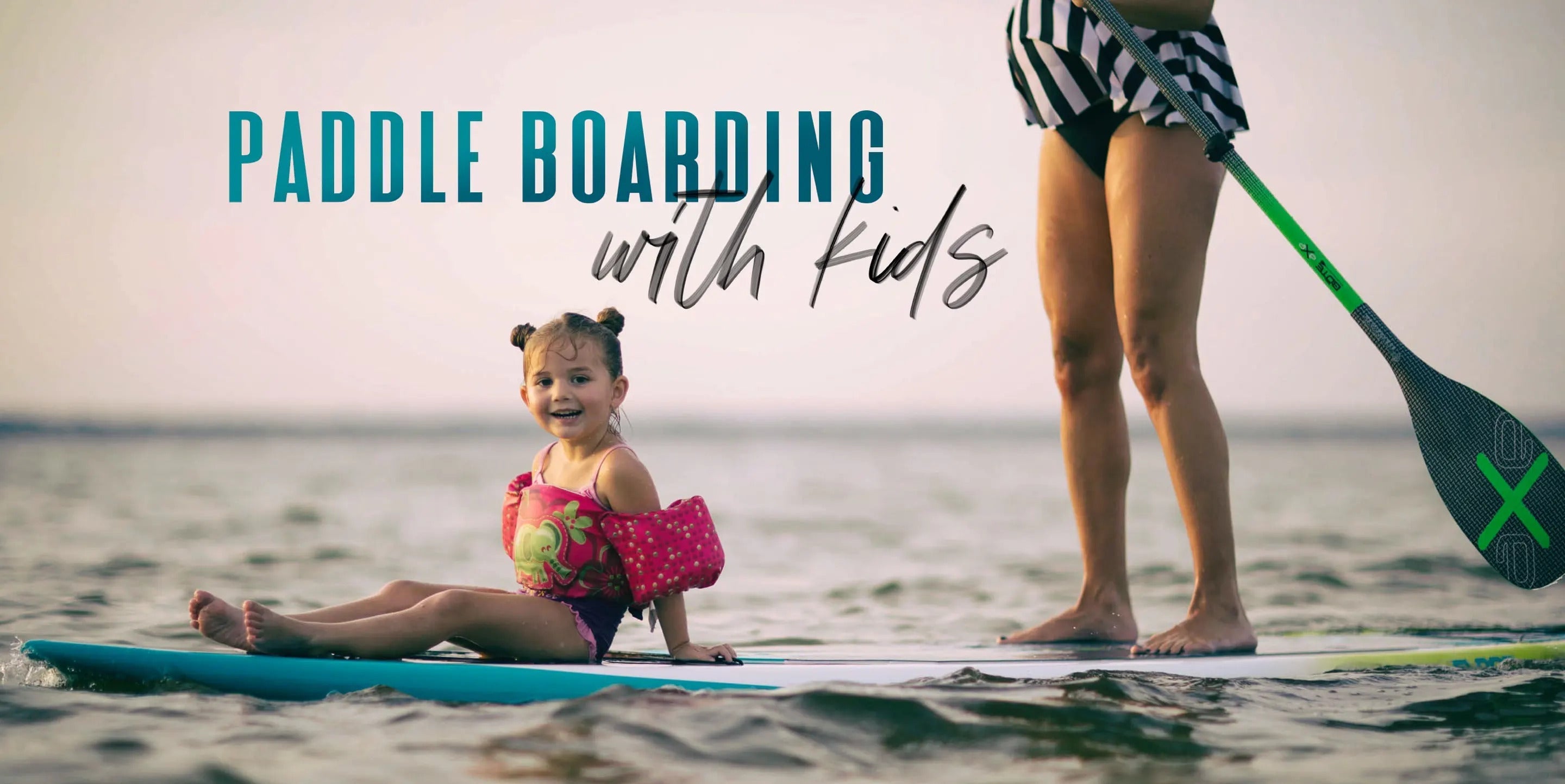 A mother paddling with her child sitting on the front of her paddle board wearing a pink and green floaty vest with the words "Paddle boarding with kids" overlayed