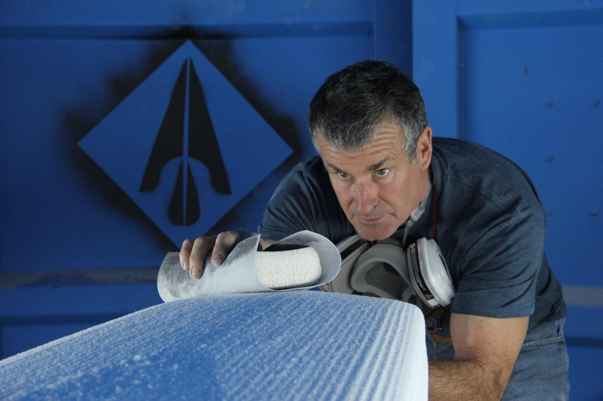 Man sanding a blue board indoors while wearing a respirator mask, with a geometric logo on the wall.