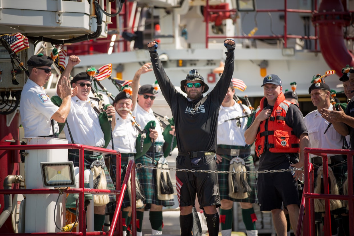 Man raising his arms in celebration on a ship deck while crew in kilts play bagpipes behind him.