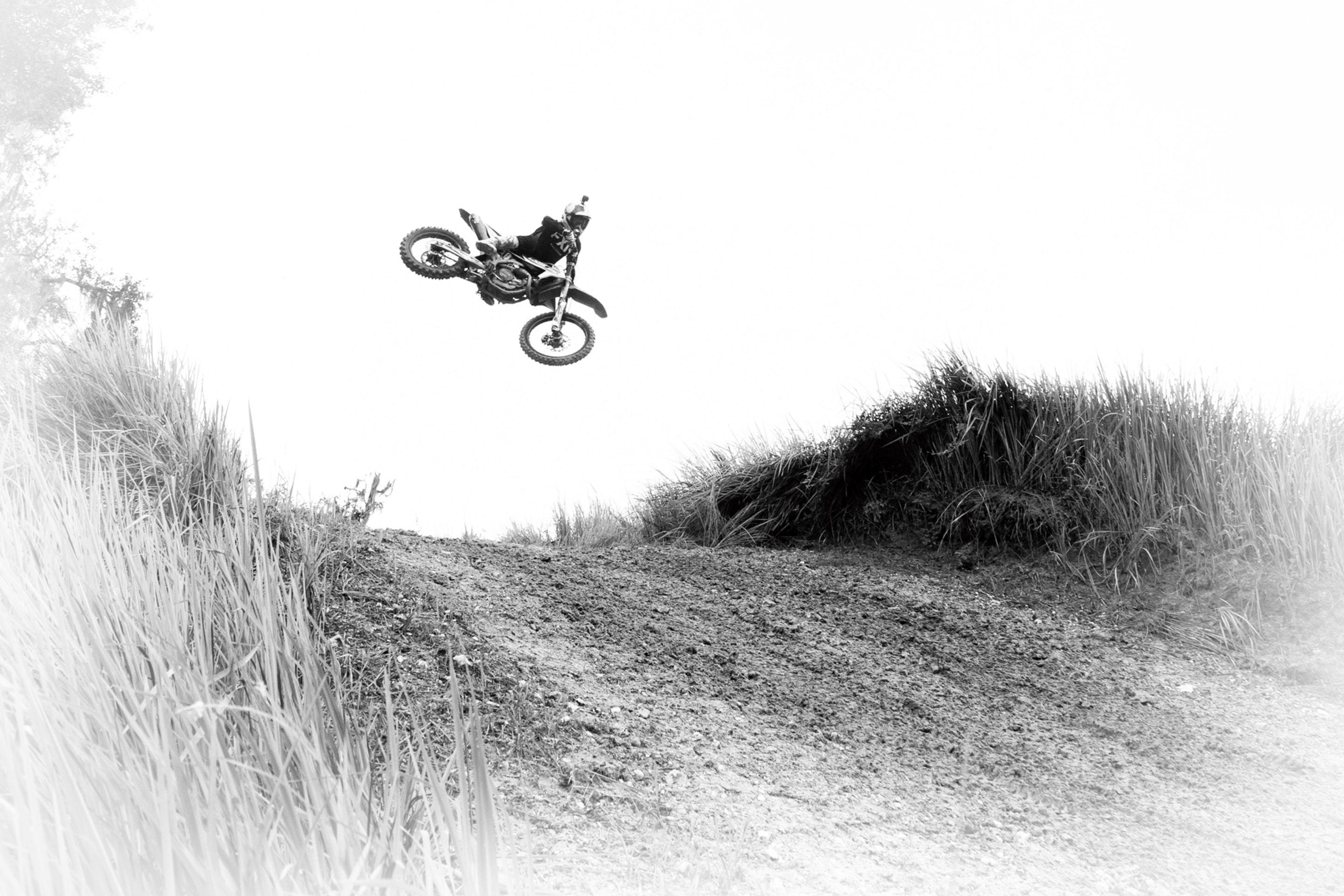 Black and white photo of a motocross rider performing a high jump over a dirt trail surrounded by tall grass.