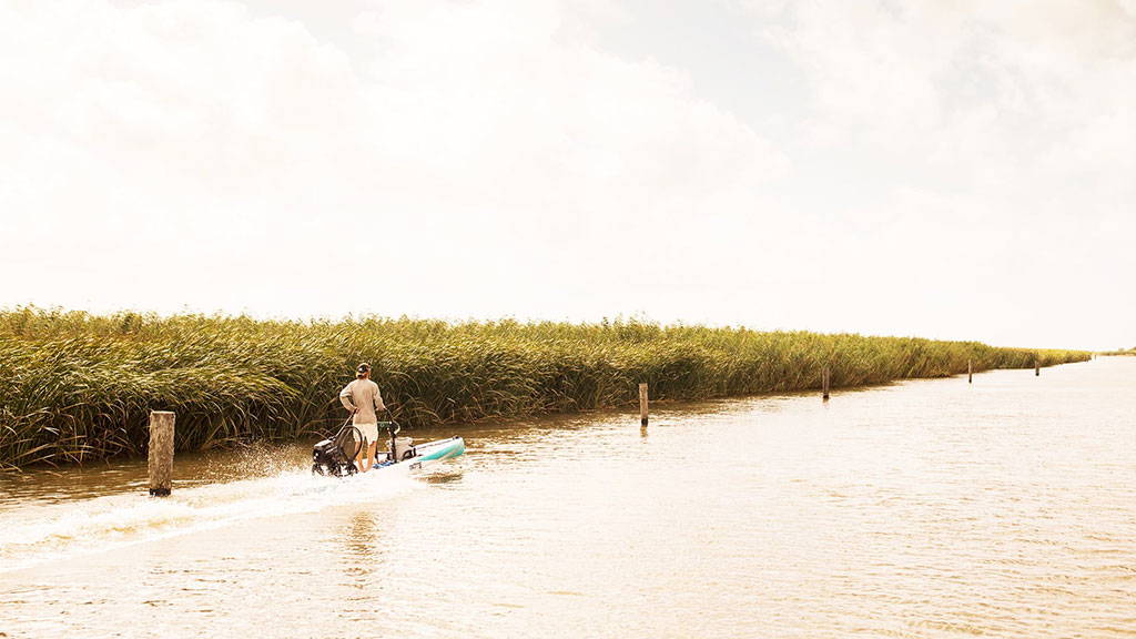 A person rides a motorized stand-up paddle board along a narrow marsh channel lined with tall reeds.
