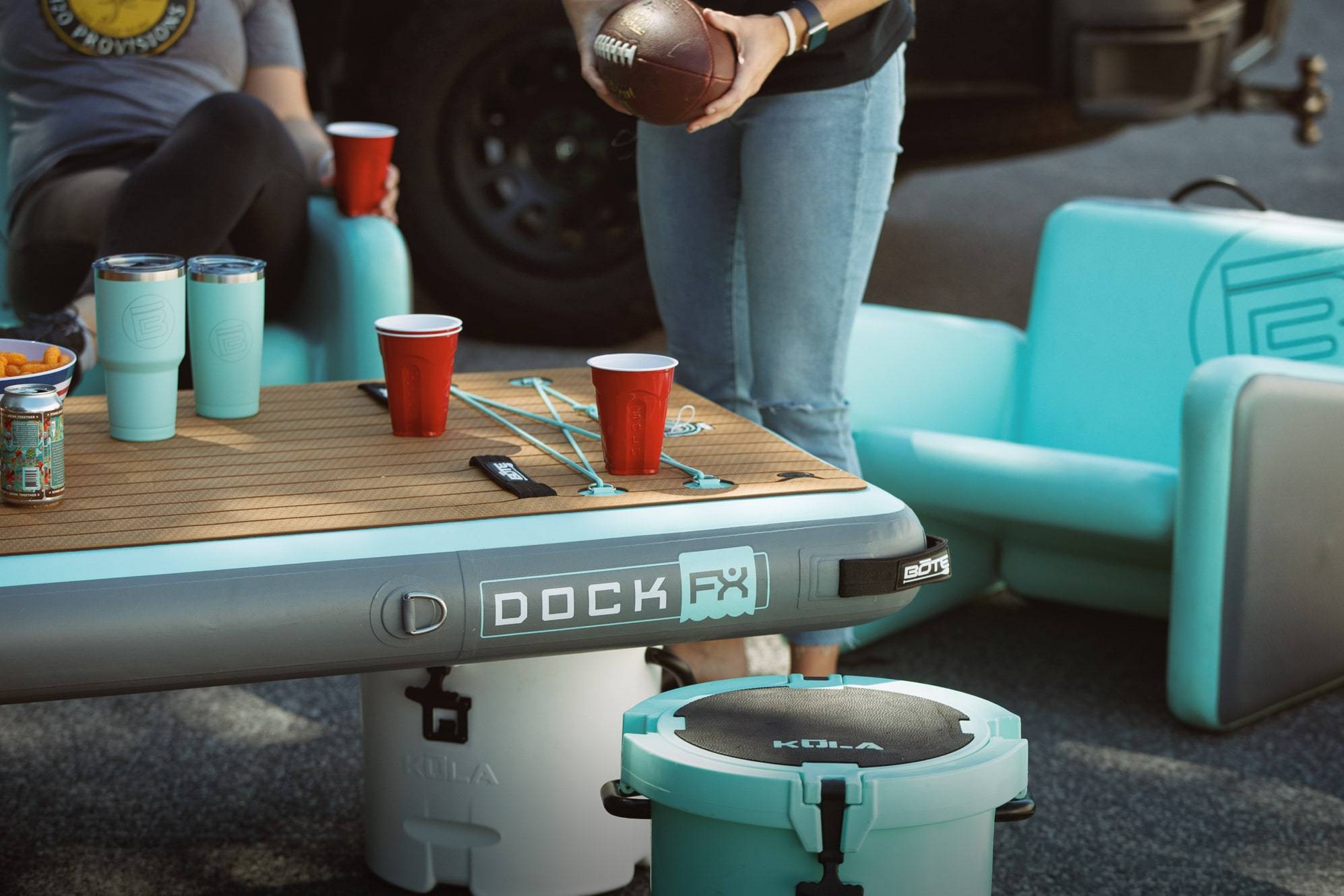 Friends tailgating with teal inflatable Dock FX table, KULA cooler, AeroChair seats, and drinks.