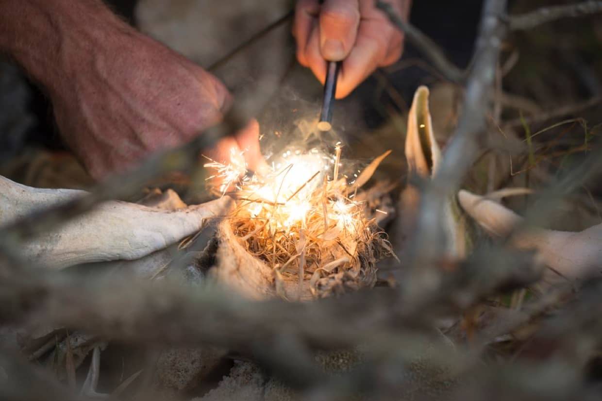 Close-up of hands using a fire starter to ignite dry tinder in a small nest of twigs.