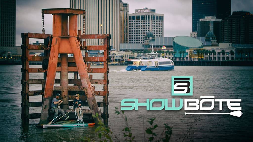 Two people fishing from paddle boards beside a rusted pier with city skyline and text reading “ShowBote.”