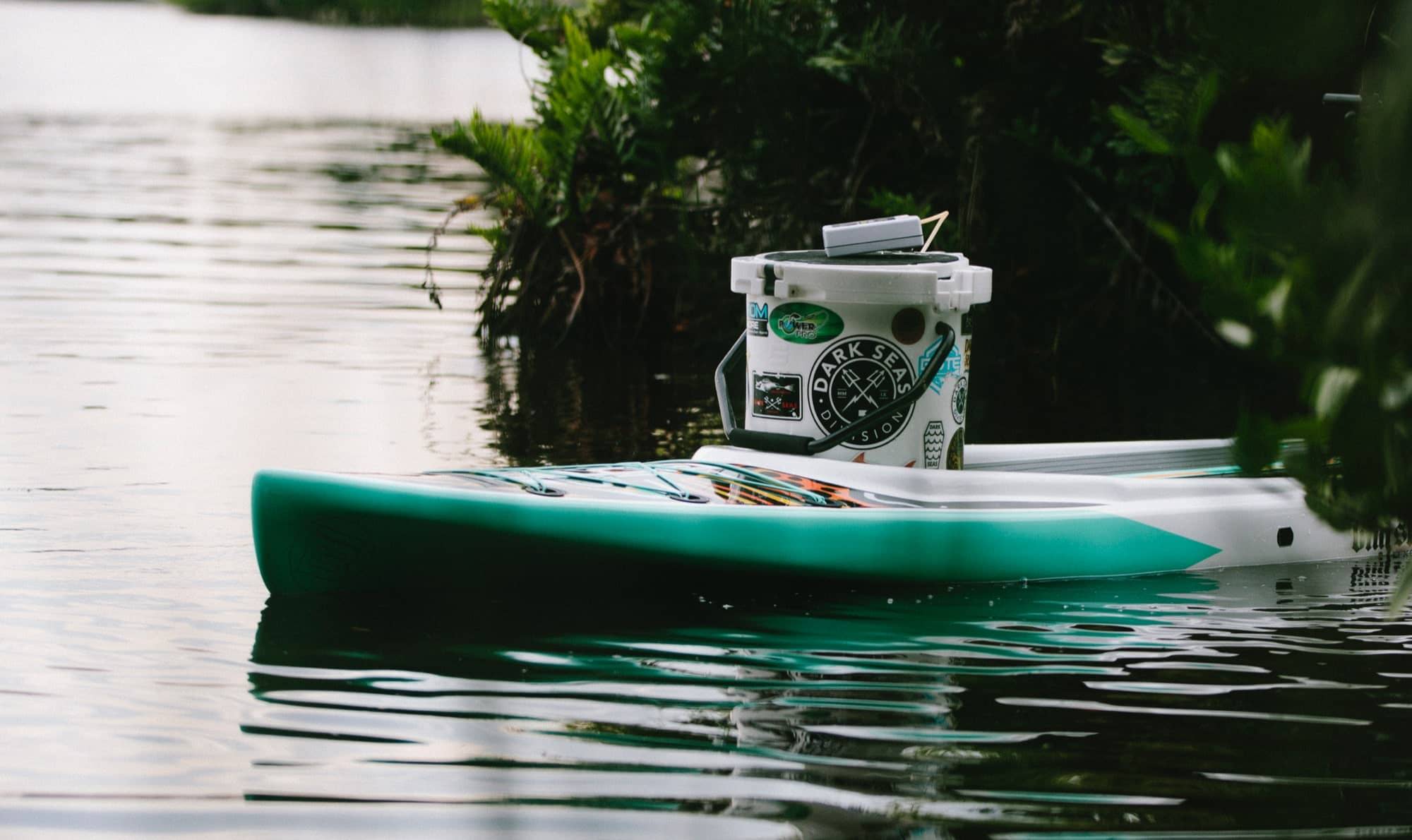 Board floating beside leafy shoreline with a sticker-covered cooler secured on the deck.