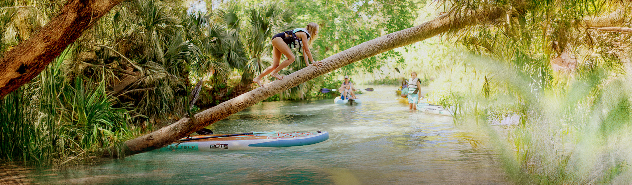 People paddleboarding on a river with a person climbing a fallen tree branch.