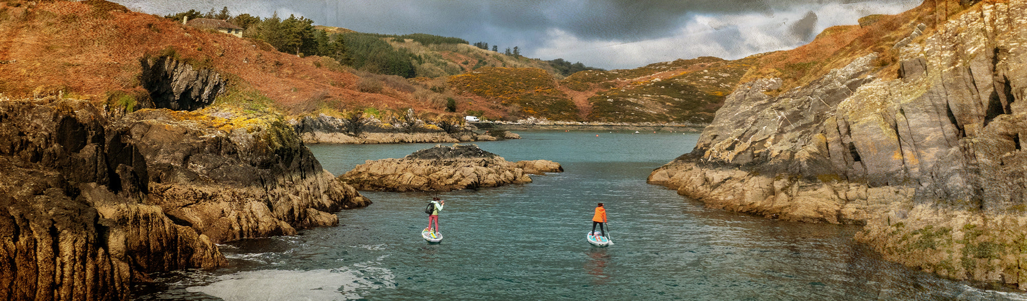 Two people paddleboarding in a coastal area with rocky cliffs and a cloudy sky.