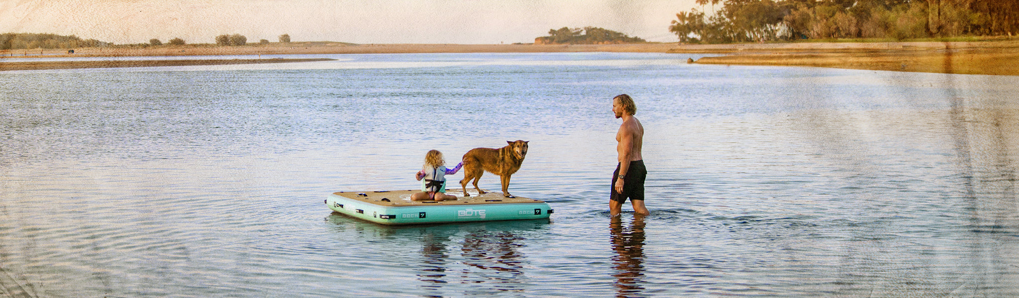 Man and two children on a small raft in a lake with a dog, surrounded by a natural landscape.