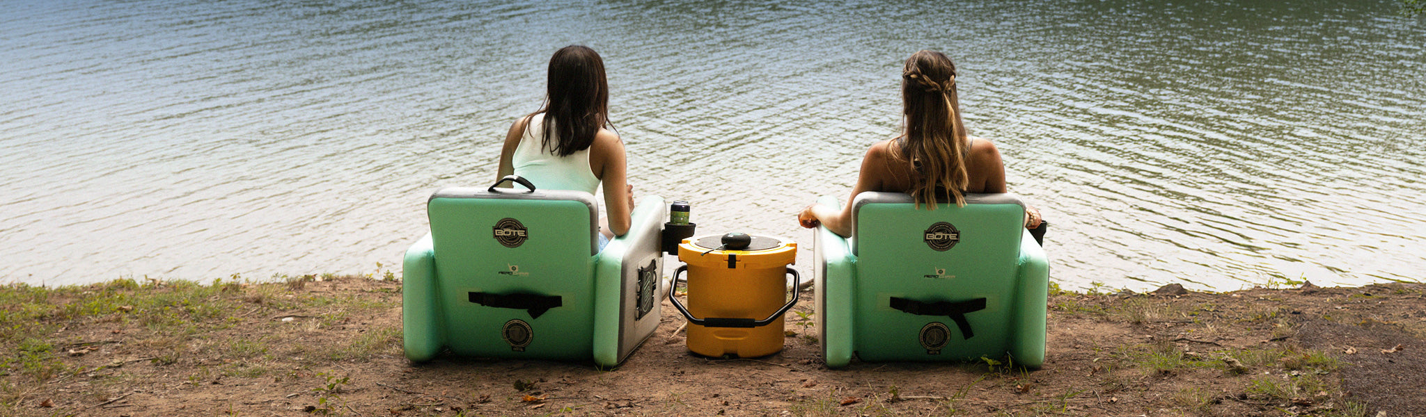 Two people sitting in green chairs by a lake with a yellow cooler between them.