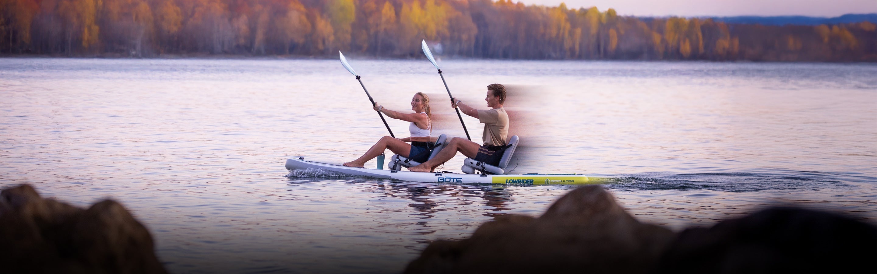 Two people sitting and paddling a paddle board on a lake with trees and mountains in the background