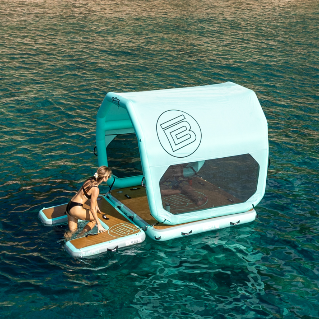Turquoise inflatable shade tent with gray mesh panels attached to a floating dock with a women climbing up the floating stairs to relax in the shade on the water.