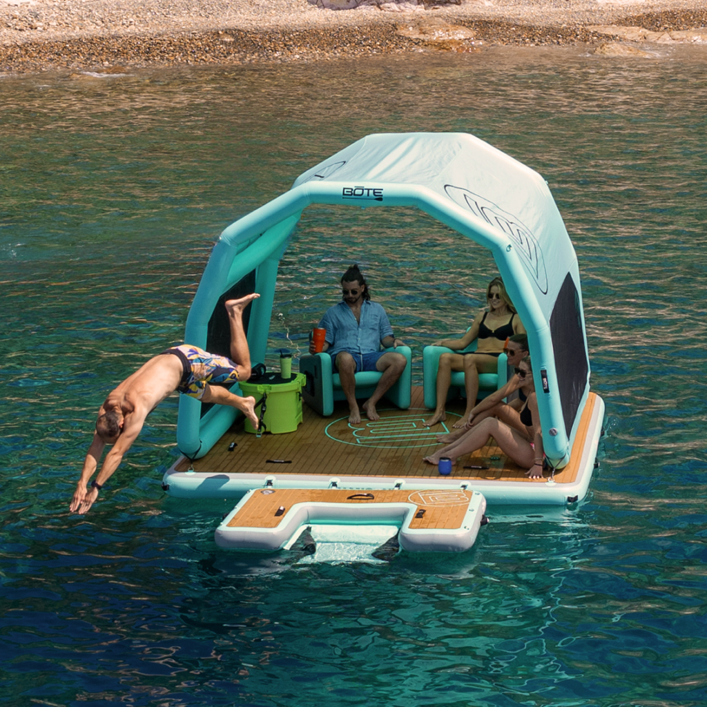 Inflatable floating platform with inflatable shade canopy attached. 4 people are lounging in the shade while one is diving off the platform.