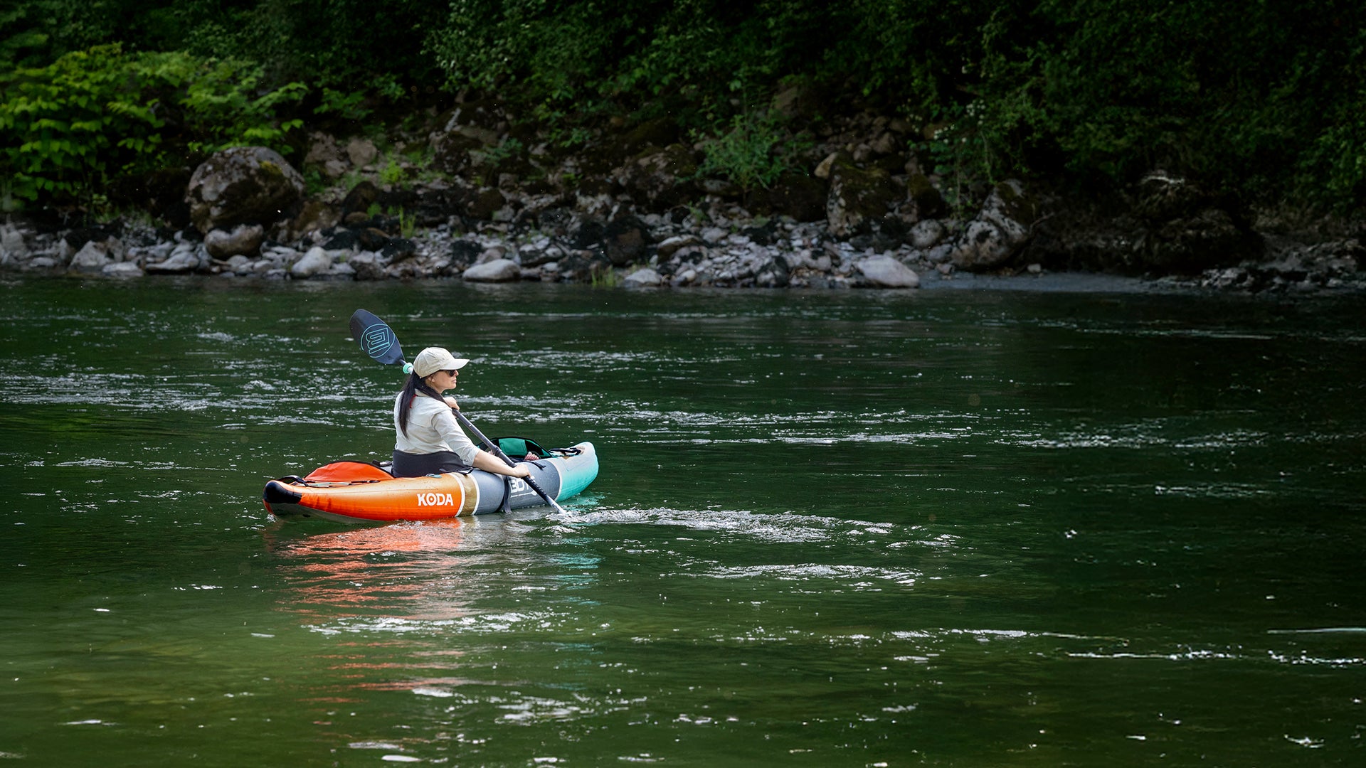 Woman paddling orange and teal Koda inflatable kayak on calm green river near rocky shore.
