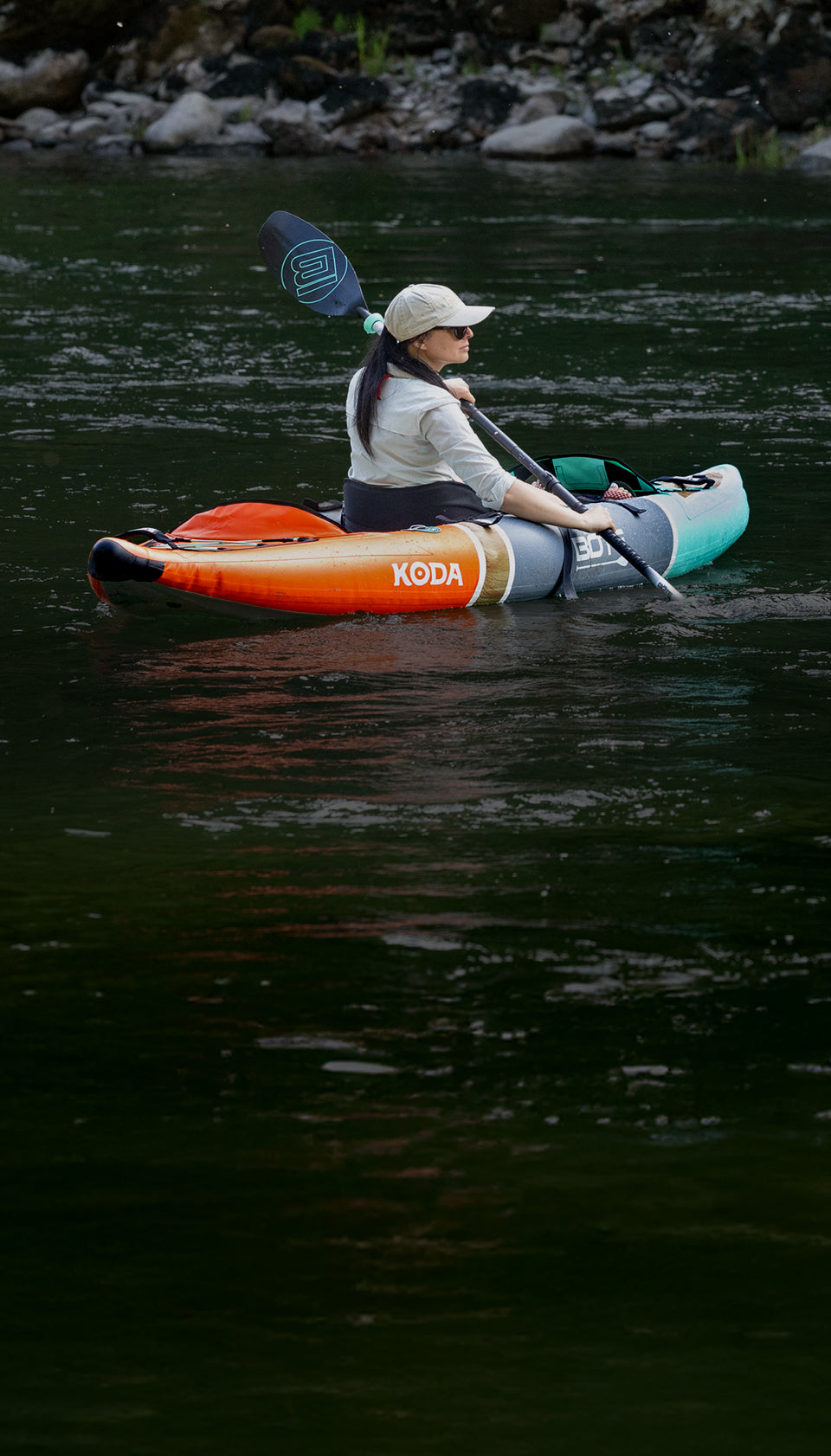 Woman paddling orange and teal Koda inflatable kayak on calm green river near rocky shore.