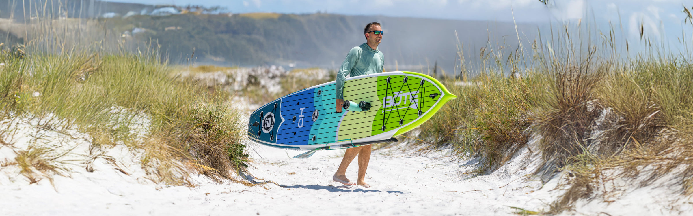 Lifestyle view of man carrying green and blue paddle board through sandy beach dunes.