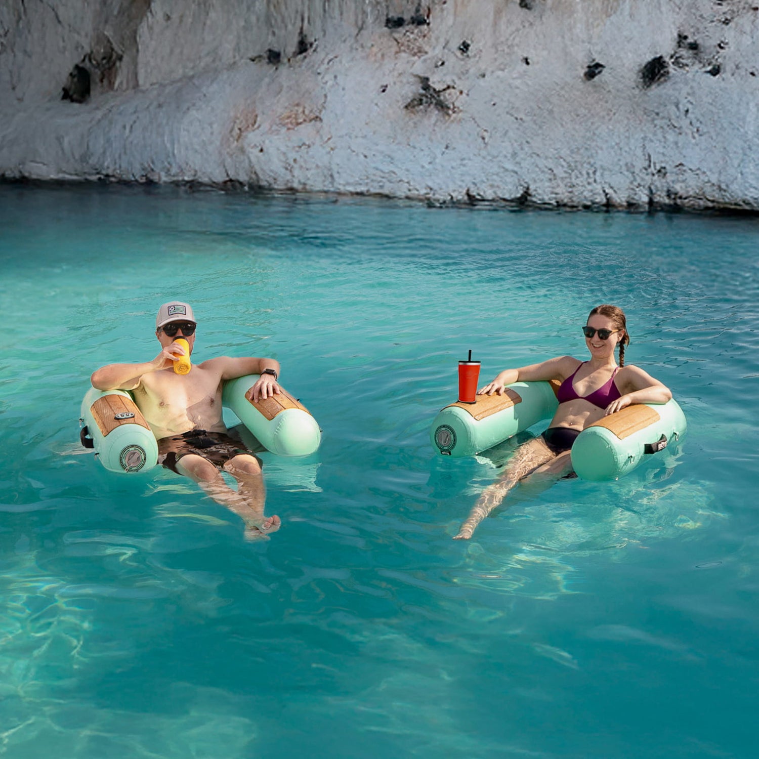 Two people relaxing on inflatable sling chairs in clear blue water with a rocky cliff in the background.