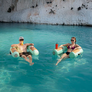 Two people relaxing on inflatable sling chairs in clear blue water with a rocky cliff in the background.