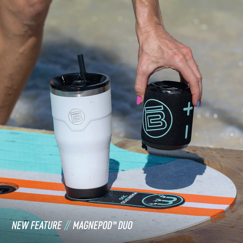 Hand placing a portable speaker next to a tumbler, both magnetically docked on a paddleboard.