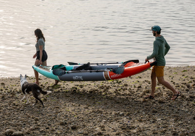 Two people carry the Koda Aero Tandem on a beach to the water as a dog walks alongside.
