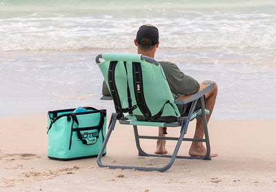 Person seated in Mule Outpost Chair at shoreline with tote bag nearby.