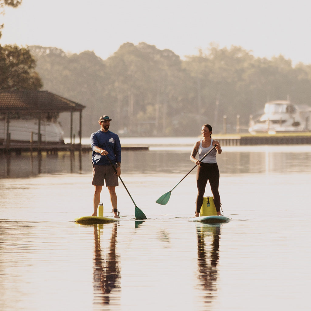 Two people paddleboarding on a calm lake with a dock and trees in the background.