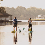 Two people paddleboarding on a calm lake with a dock and trees in the background.