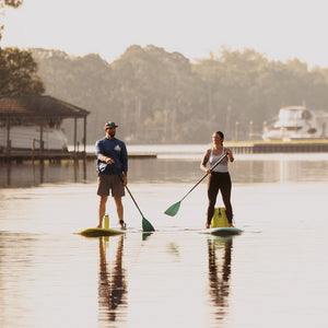 Two people paddleboarding on a calm lake with a dock and trees in the background.
