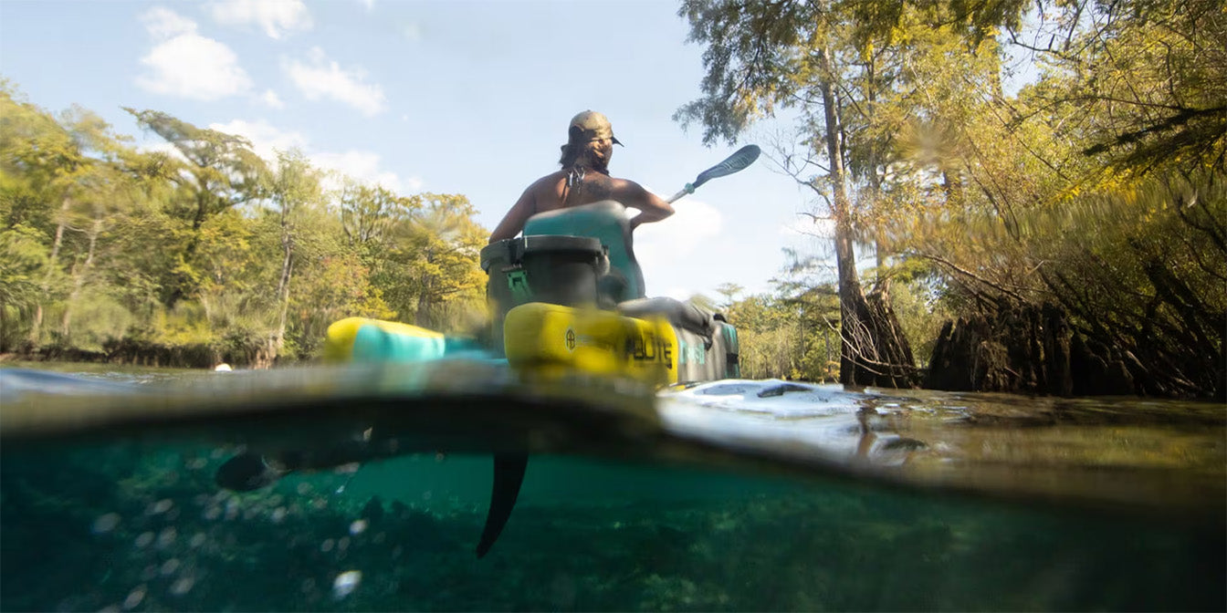 Rear view of paddler kayaking through forested waterway, half-underwater perspective.