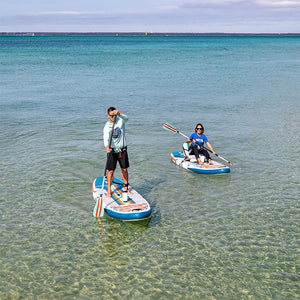 Two people paddling EasyRider Aero boards in shallow clear water near the shoreline.