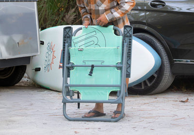Person holding folded Mule Outpost Chair near vehicle and paddle board.