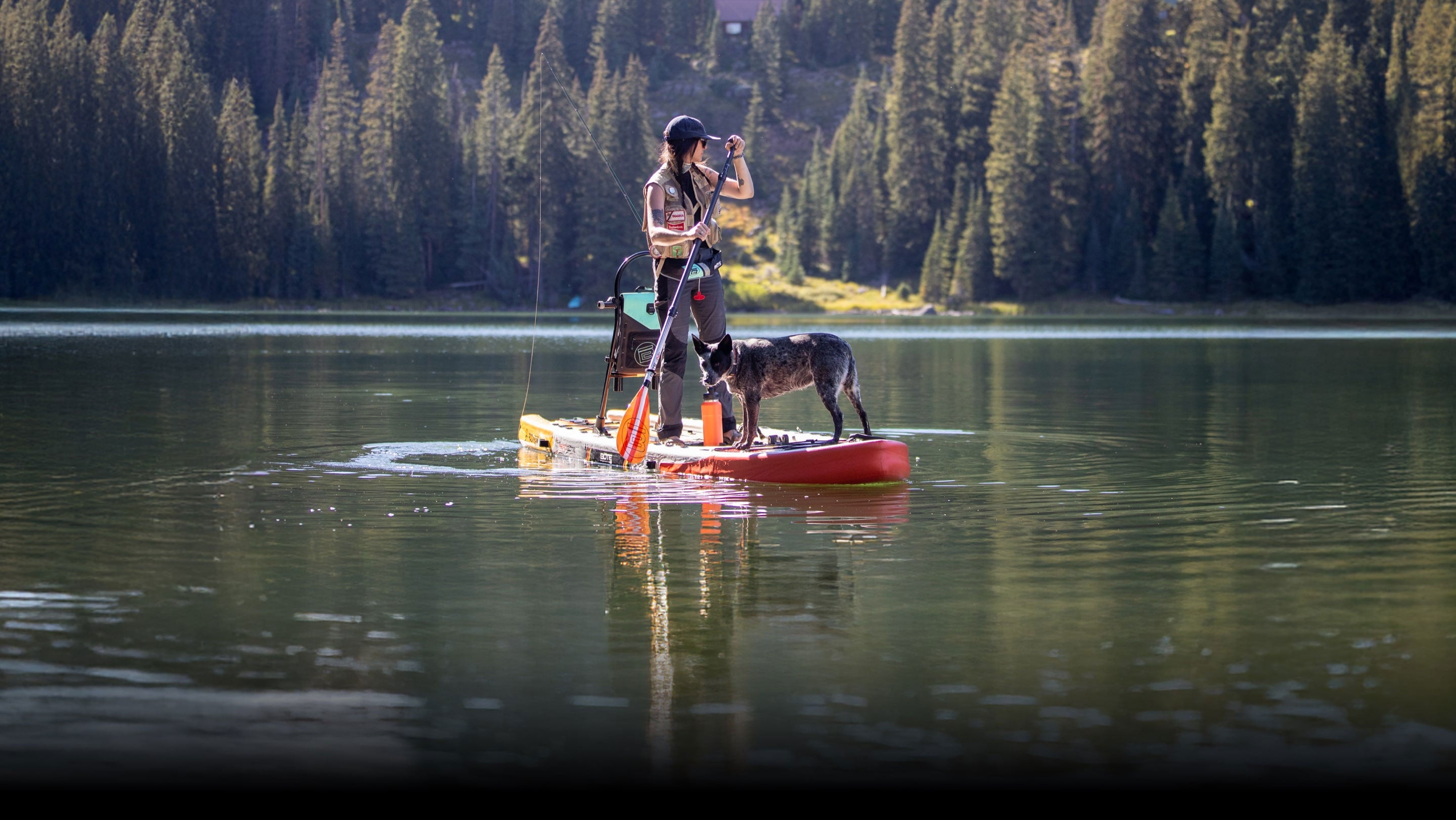 Woman paddling on a Rackham Aero with her dog across a calm lake surrounded by trees.