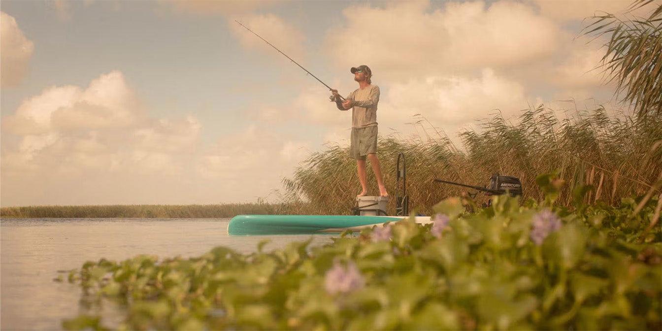 Man fishing while standing on Rover Gatorshell micro skiff near tall reeds.