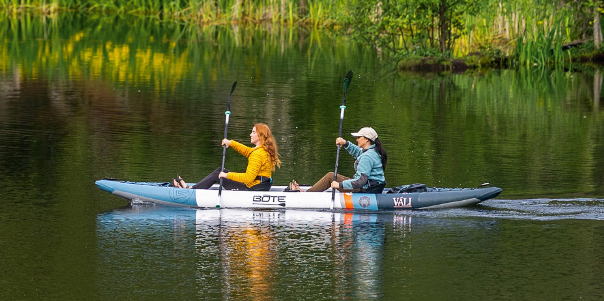 Two women paddling the Vali Aero kayak on a still green lake surrounded by trees.
