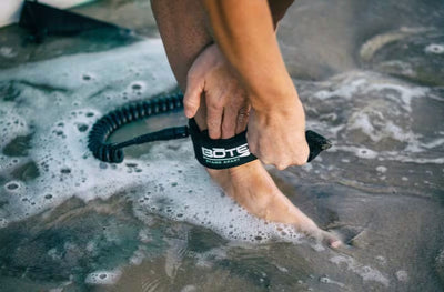 Close-up of a person securing an ankle leash labeled with the BOTE logo in shallow water.