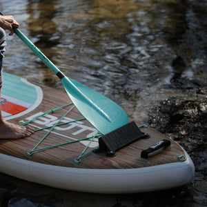 Close-up of paddle board with paddle resting on bungees near water.