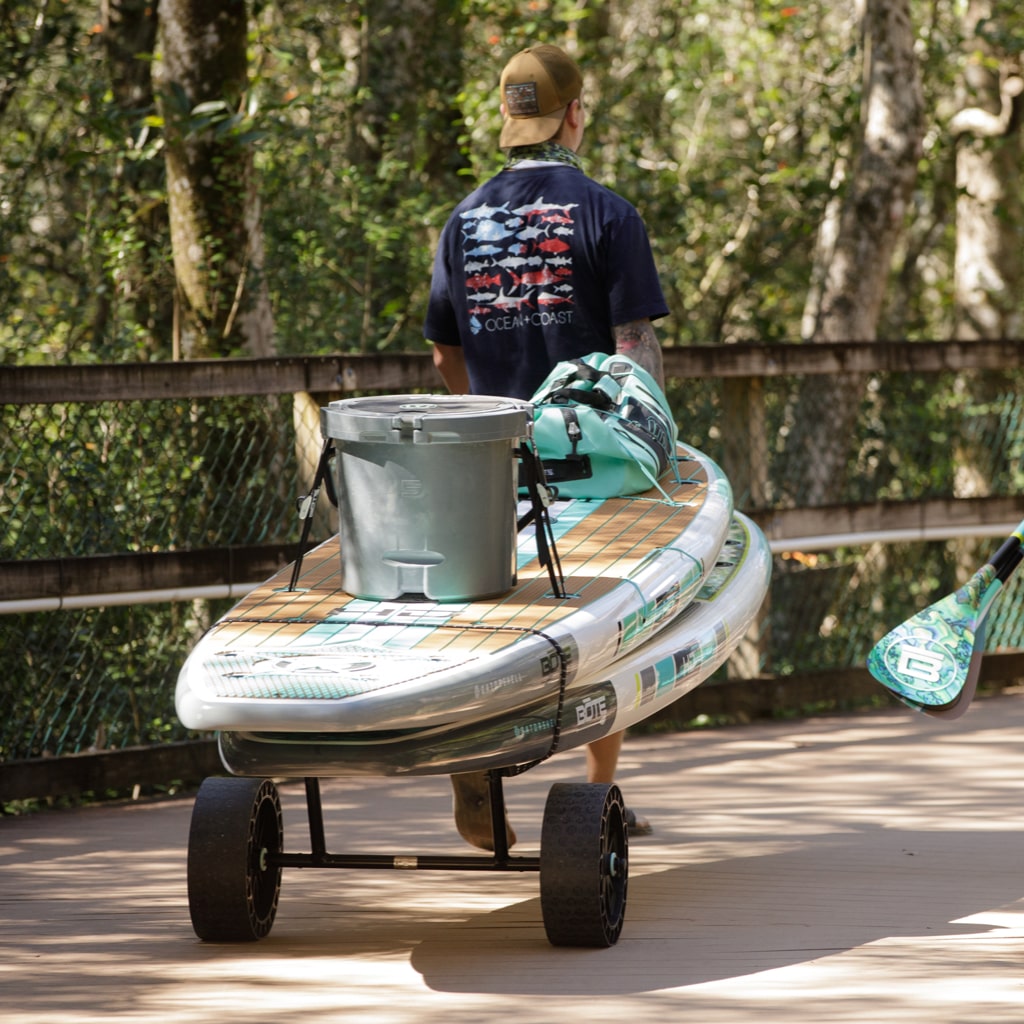Man pulling a loaded paddle board fitted with Wheel Rac along a wooden path.