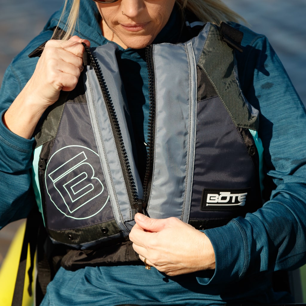Woman zipping up the BOTE Adult Paddling Foam PFD Vest while seated outdoors near water.